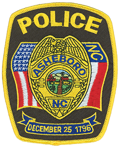 The badge of the Asheboro, North Carolina, Police Department is prominently depicted on the agency’s service patch. The Asheboro city seal is featured in the center. Within the seal is a depiction of a plank road that ran through the city in the 1700s, a prominent church from Asheboro’s early days, and a textile factory. The flags of the United States and North Carolina are shown to the left and right of the badge, respectively. The banner at the bottom displays the date on which Asheboro was chartered—Christmas Day, 1796.