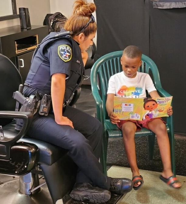 A photo of an Irving police officer reading a book with a young child in the barber shop.