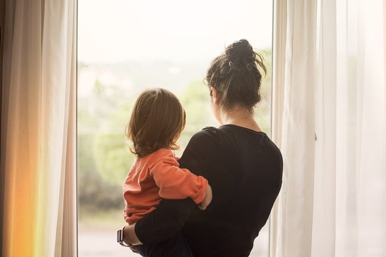 A stock image of a Mom holding her child and looking out a window.