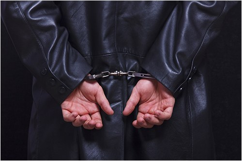 Stock image of a man in a dark jacket handcuffed behind his back.
