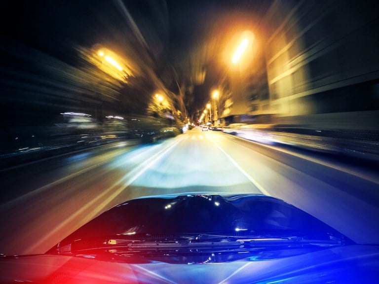 A stock image of a police car driving at night with the emergency lights on.
