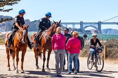 Two police officers on horses talk to citizens on a gravel path. 