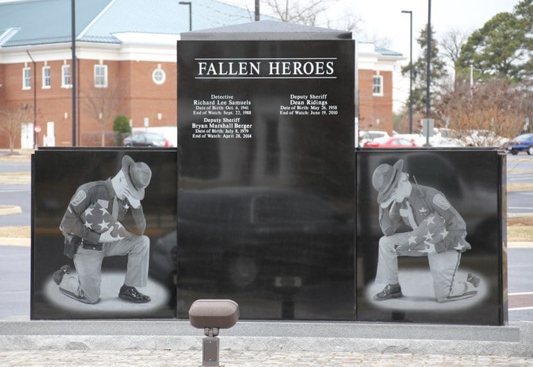 Photo of the Spotsylvania County Sheriff’s Office Law Enforcement Memorial. The photo depicts the side of the memorial which includes images of a female and male officer kneeling while holding a folded American flag and the engraved names of those who have died in the line of duty between the two officers.