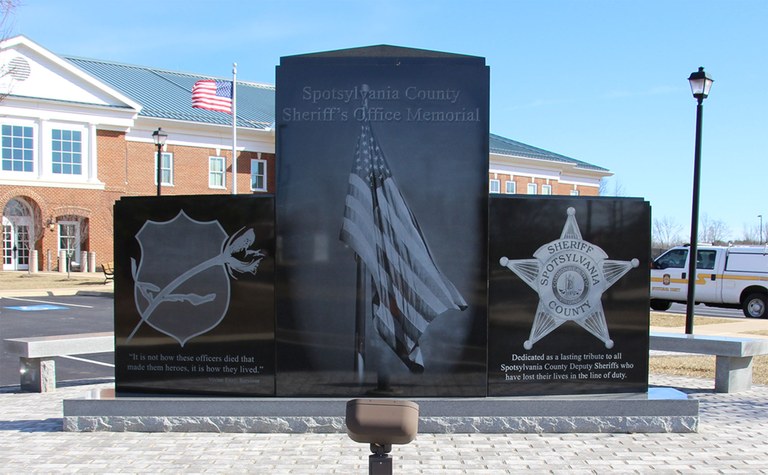 Photo of the front of the Spotsylvania County Sheriff’s Office Law Enforcement Memorial which includes the symbolic blue badge and rose, the American flag, and the badge of the Spotsylvania County Sheriff's Office.