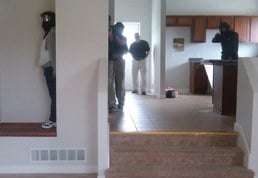 Students move toward the living room while an instructor watches. The house presents multiple danger areas for students to address even though there is no furniture.